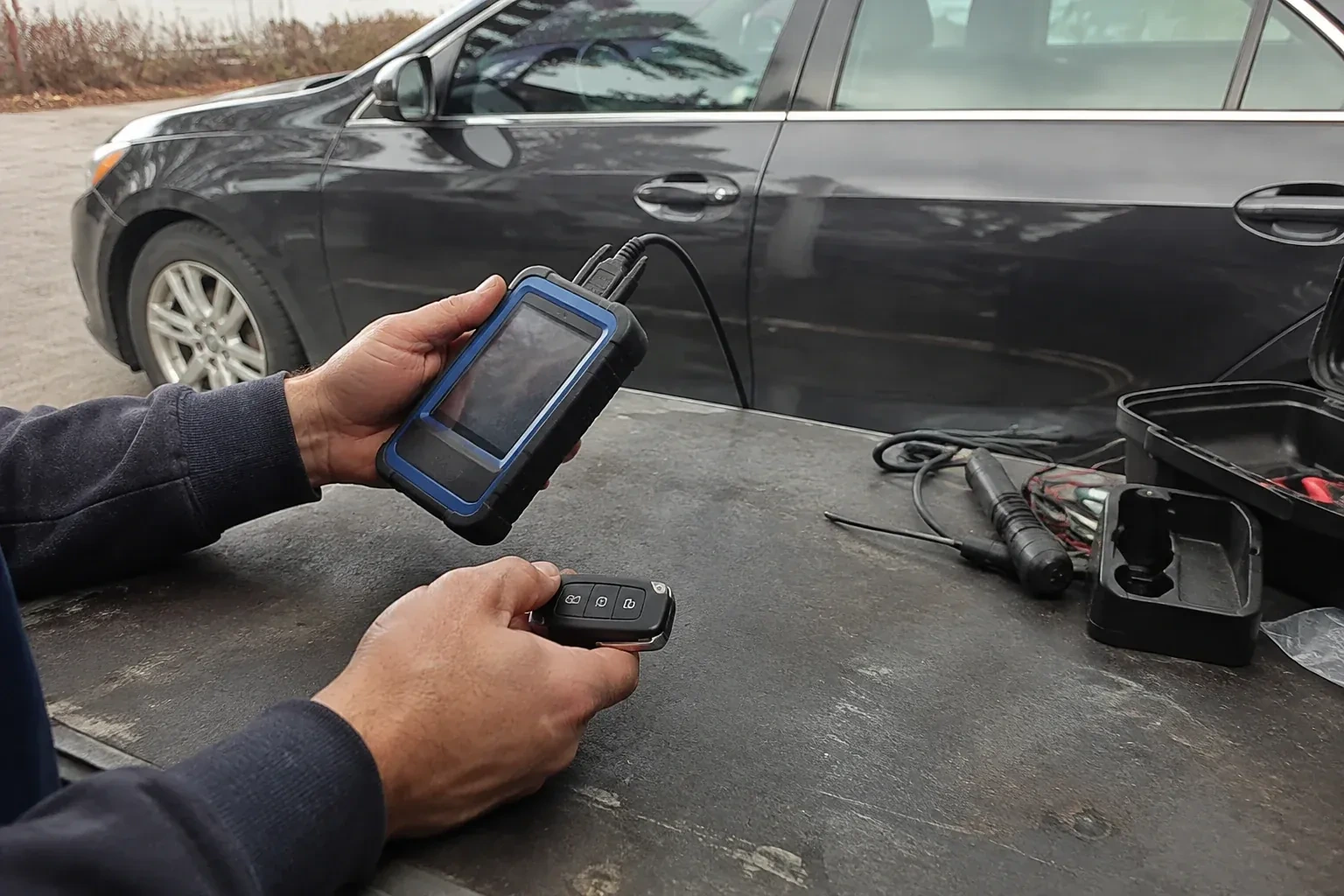 Hands holding a blue-edged electronic device and car-key fob at a worktable beside a parked car. — Transponder Key Programming, Vaughan