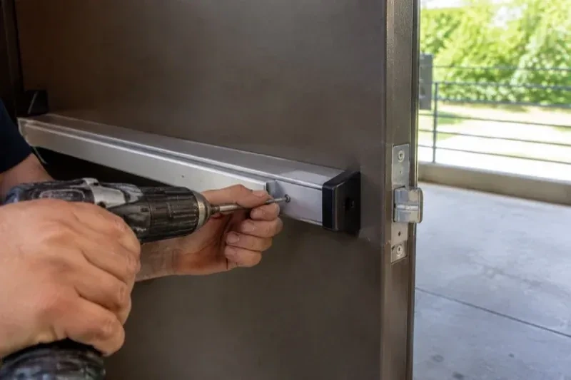 Hands using a power driver at the end of a horizontal push bar on an open metal door. — Panic & Exit Hardware, Vaughan
